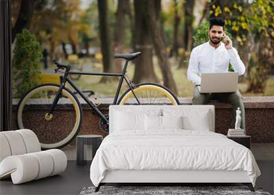 Charming young indian businessman talking on mobile phone while working on laptop computer sitting outdoors with bicycle Wall mural